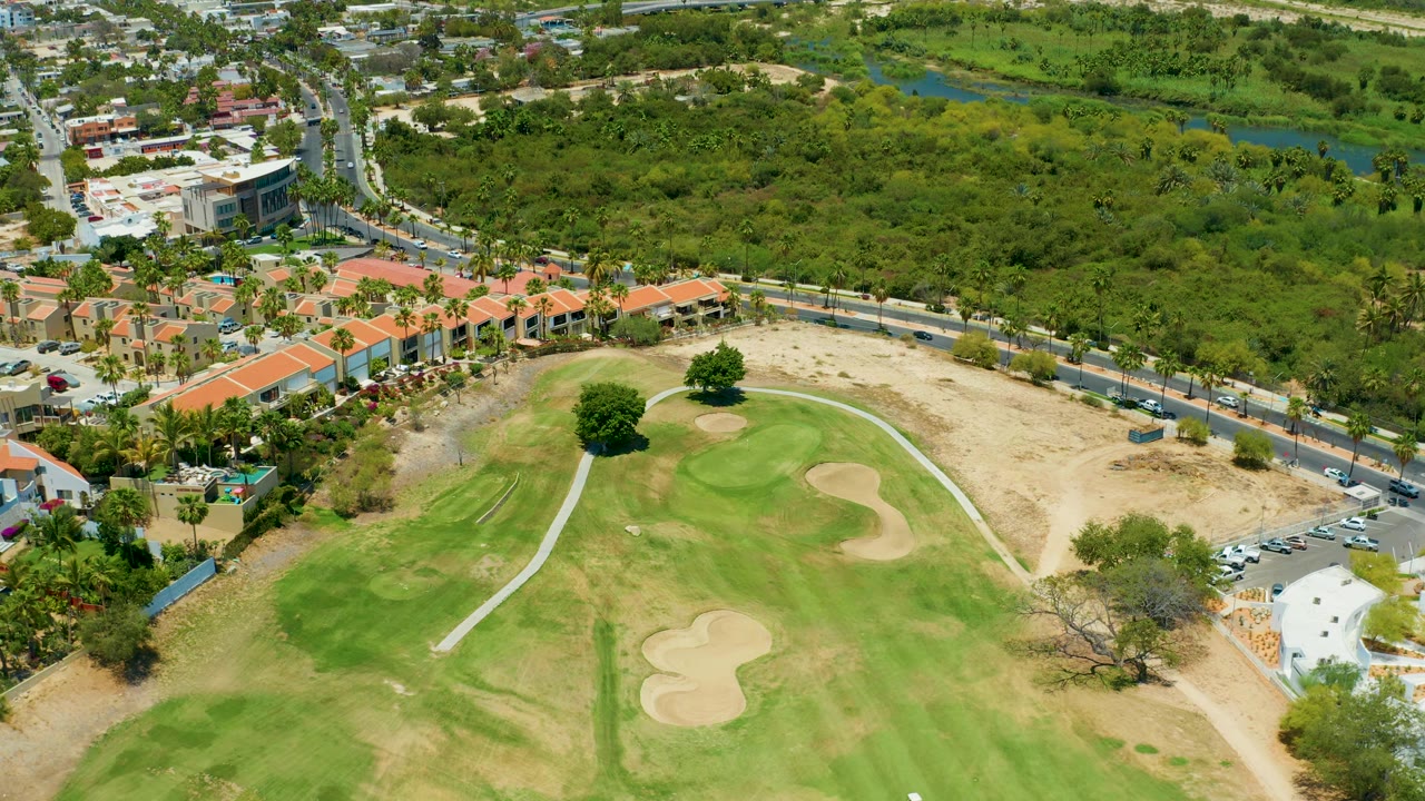 San Jose Del Cabo aerial view showing the right side of the golf course, tan sand bunkers, orange-roofed homes, a road with small vehicles, and broad green vegetation beyond, with no visible people.