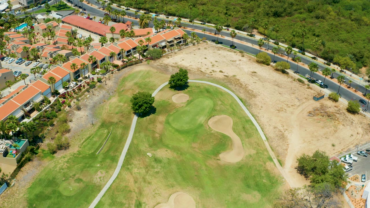 A high aerial frame in San Jose Del Cabo shows a green golf course with sand traps, a pale path, dry ground, a road, red-roofed buildings, parked cars, palm trees, and no visible people.