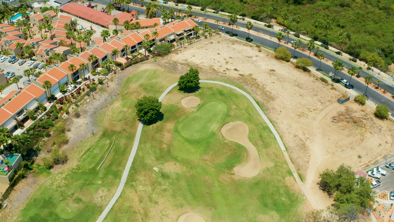 A later aerial frame in San Jose Del Cabo shows the golf course shifted slightly in the view, with sand traps, a pale path, dry open ground, the road, vehicles, red roofs, palm trees, and no visible people.