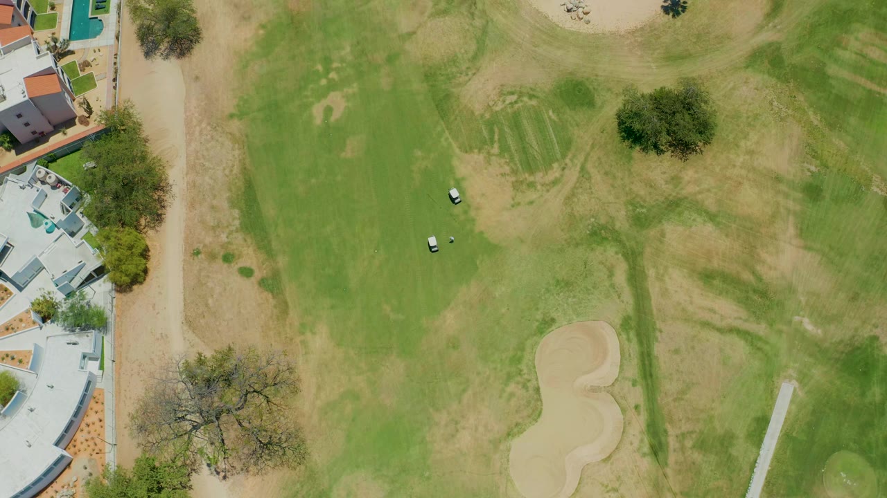 Overhead view of a San Jose Del Cabo golf course with two white carts on green and dry fairway grass, sand traps to the right, buildings along the left edge, and no visible people.
