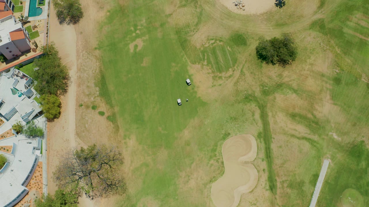 Overhead frame of a San Jose del Cabo golf course showing two small golf carts on green fairway grass, pale sand traps, trees, buildings at left, and no visible people.