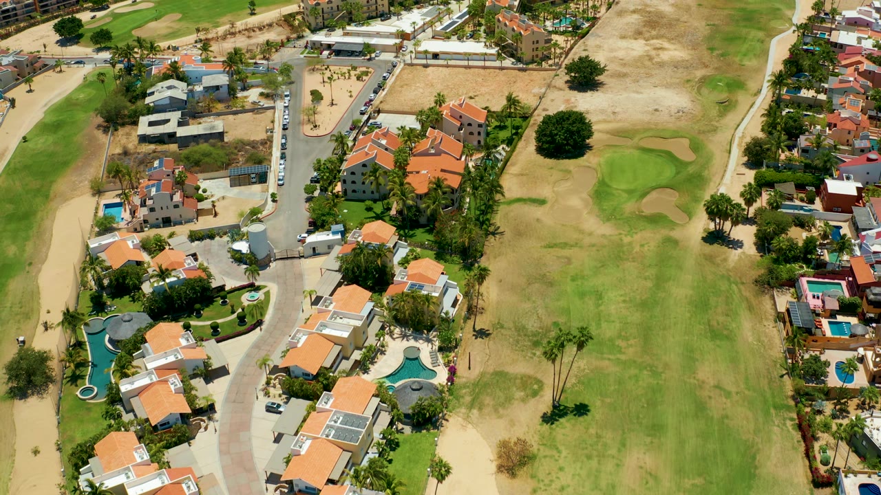 San Jose Del Cabo aerial frame showing residential blocks, orange roofs, small pools, a pale road, golf bunkers, dry turf, and no visible people.