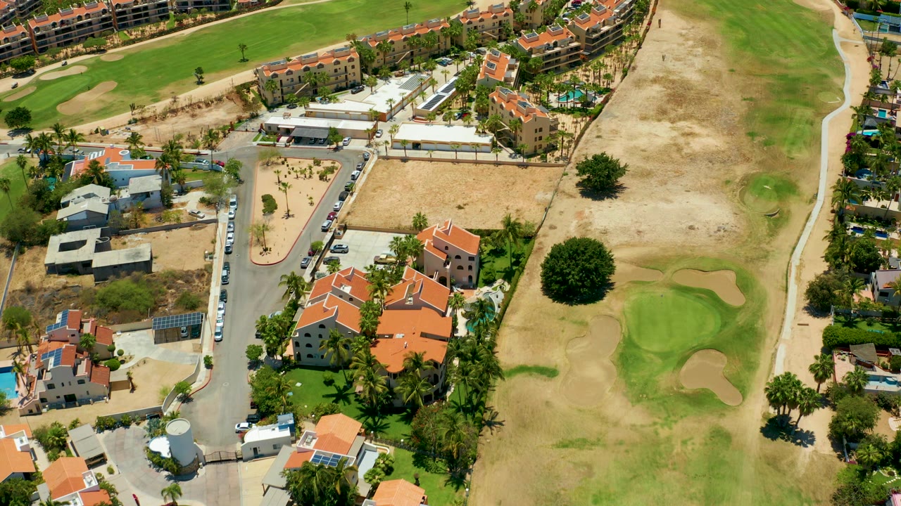 Aerial frame from San Jose Del Cabo with residential buildings at lower left, a curved road with cars, tan open ground, fairway patches, and no visible people.