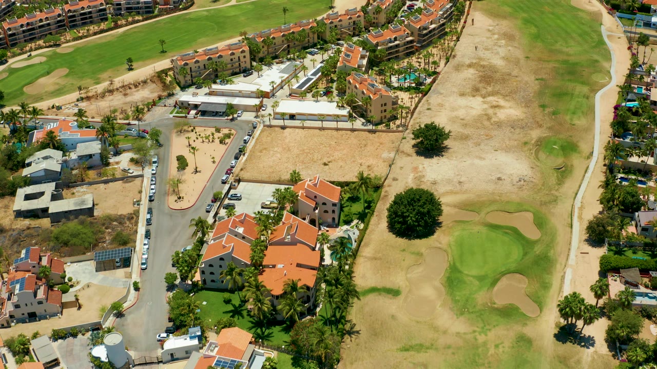 Aerial frame from San Jose Del Cabo showing homes with orange roofs, a narrow road lined with cars, a large dry course area, green fairway sections, and no visible people.