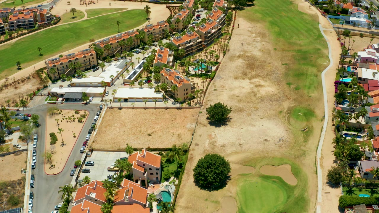Aerial frame over San Jose del Cabo showing red-roofed residential buildings, a parking area, dry ground, green fairway, sand traps, roads, trees, and no visible people.