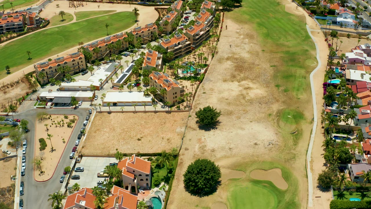 High oblique frame over San Jose del Cabo showing a row of red-roofed buildings, adjacent homes, dry golf course rough, a green putting area, a sand trap, roads, and no visible people.