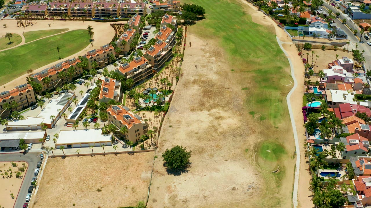 Aerial view in San Jose Del Cabo showing orange-roofed residential buildings, palm landscaping, a road, and a green-brown field at the start of the clip.