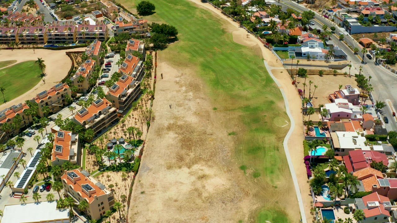 Aerial frame over San Jose del Cabo showing red-roofed residences, pools, palms, a road, and a large patchy grassy field with no visible people.