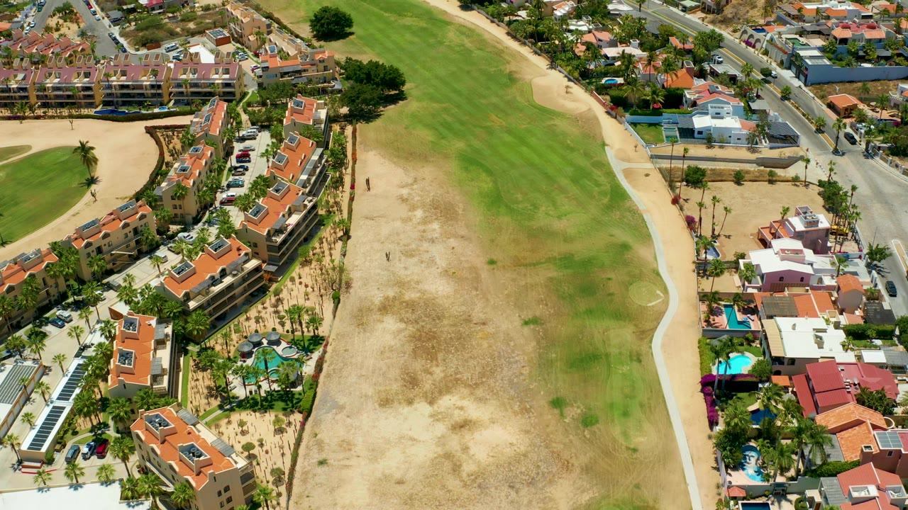 Aerial view in San Jose del Cabo with a curved row of red-roofed homes beside the open field, pools and palms below, and a paved road crossing the upper frame.