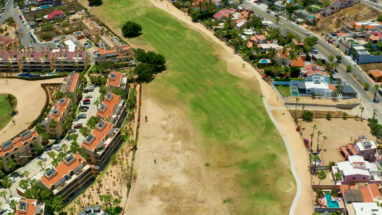 Aerial frame in San Jose Del Cabo with the field centered, a pale sandy path curving along the right side, red-roof buildings below, and cars on the road, with no visible people.