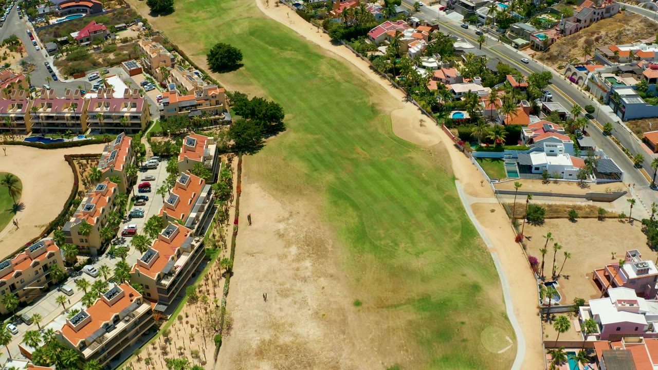 Aerial frame over San Jose Del Cabo showing the grassy field stretching upward, sandy margins, red tile roofs, a road with vehicles, and small pools near homes, with no visible people.