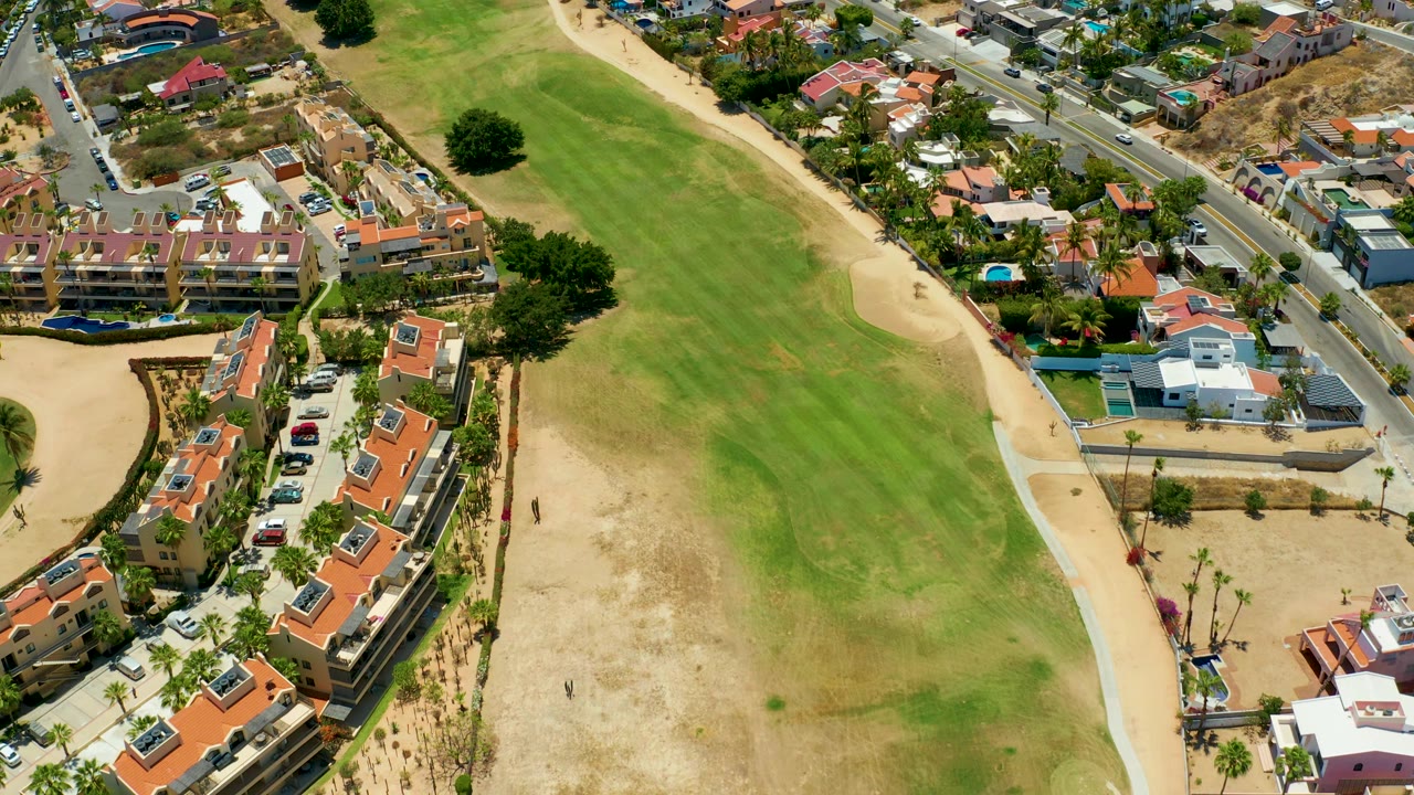 Aerial residential coastline view with a large field, sandy edge, apartments, road, and cars.