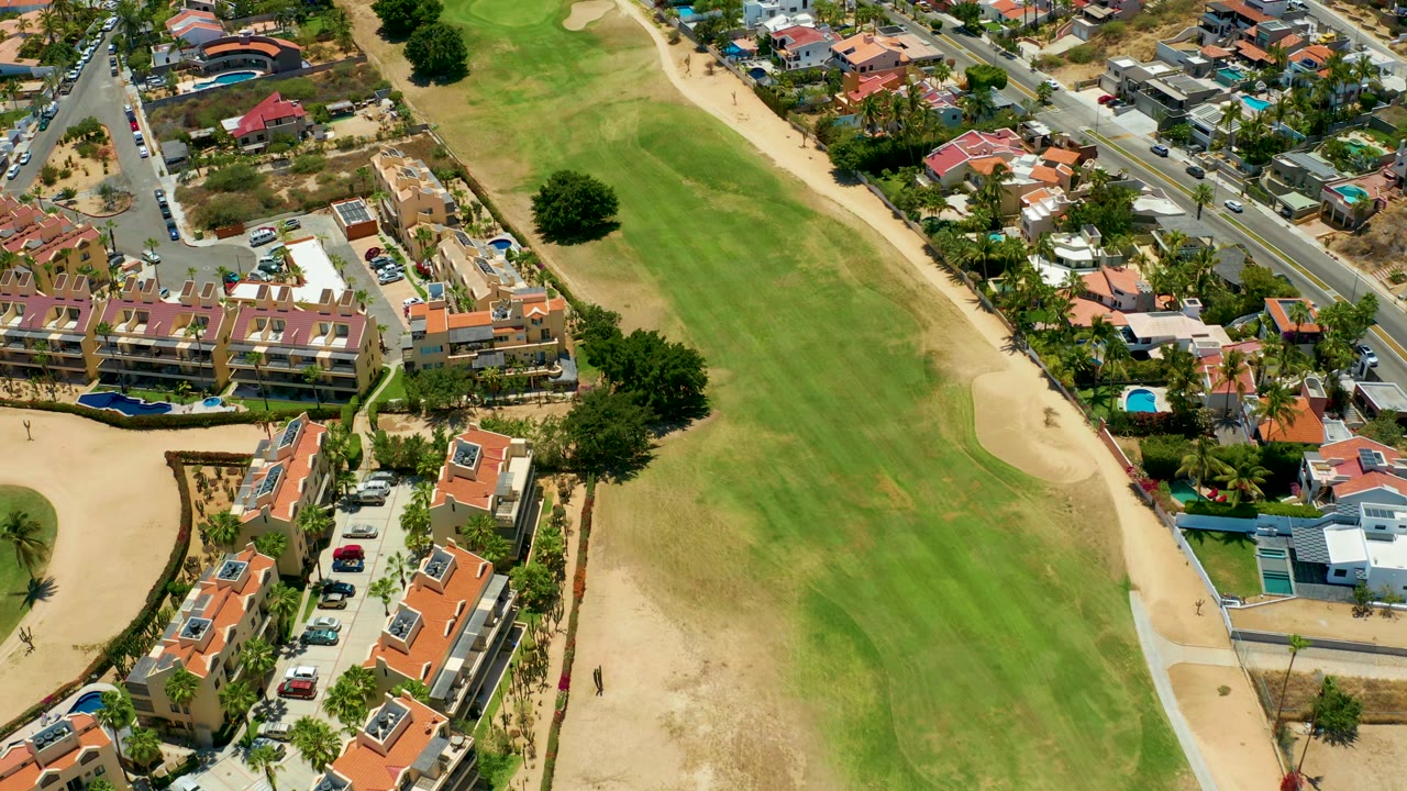 Aerial frame in San Jose Del Cabo with the fairway running through the center, houses and pools along both sides, road traffic near the top, and no visible people.
