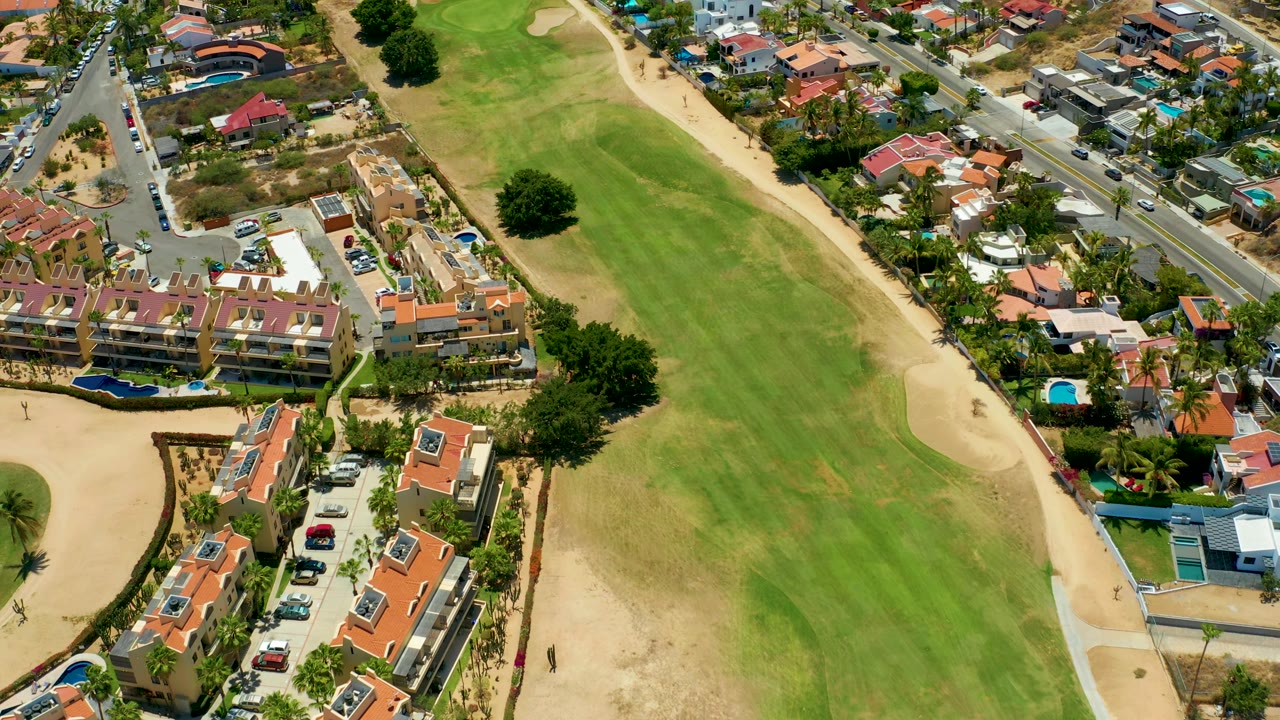 San Jose Del Cabo aerial view with broad green fairway in the lower frame, tan bunker edges, clustered homes, parked vehicles, and no visible people.