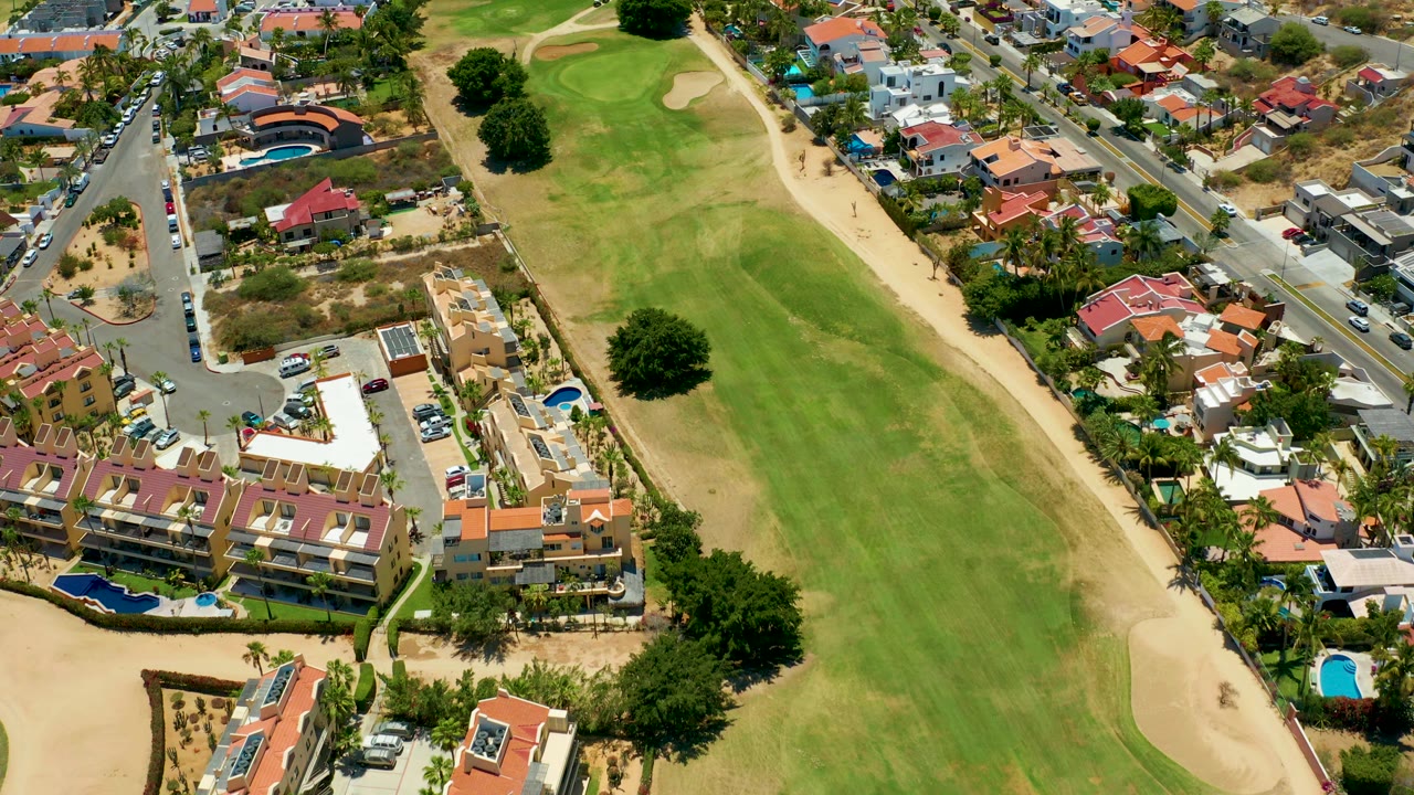 Opening aerial frame over San Jose Del Cabo showing a green golf fairway, sand along the right edge, nearby homes, pools, streets, cars, and no visible people.