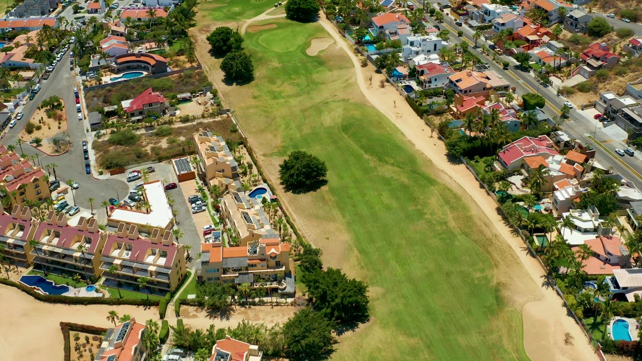 Mid-run aerial view over San Jose Del Cabo with the green fairway shifting across frame, a curved street junction, red roofs, parked cars, pools, and no visible people.