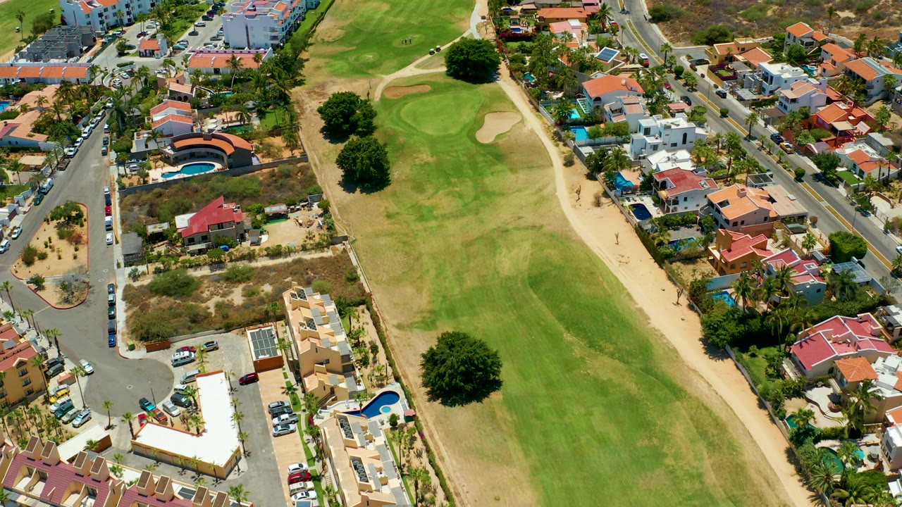 Aerial frame from San Jose Del Cabo showing a central green golf fairway bordered by tan paths, houses, pools, roads, and dry lots with no visible people.