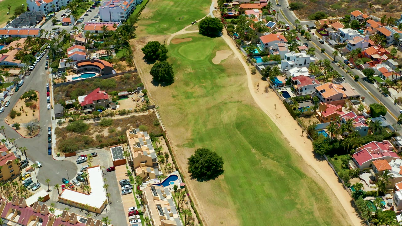 Aerial view in San Jose Del Cabo with the golf green and bunker near center, a curved residential road, parked cars, pools, and no visible people.