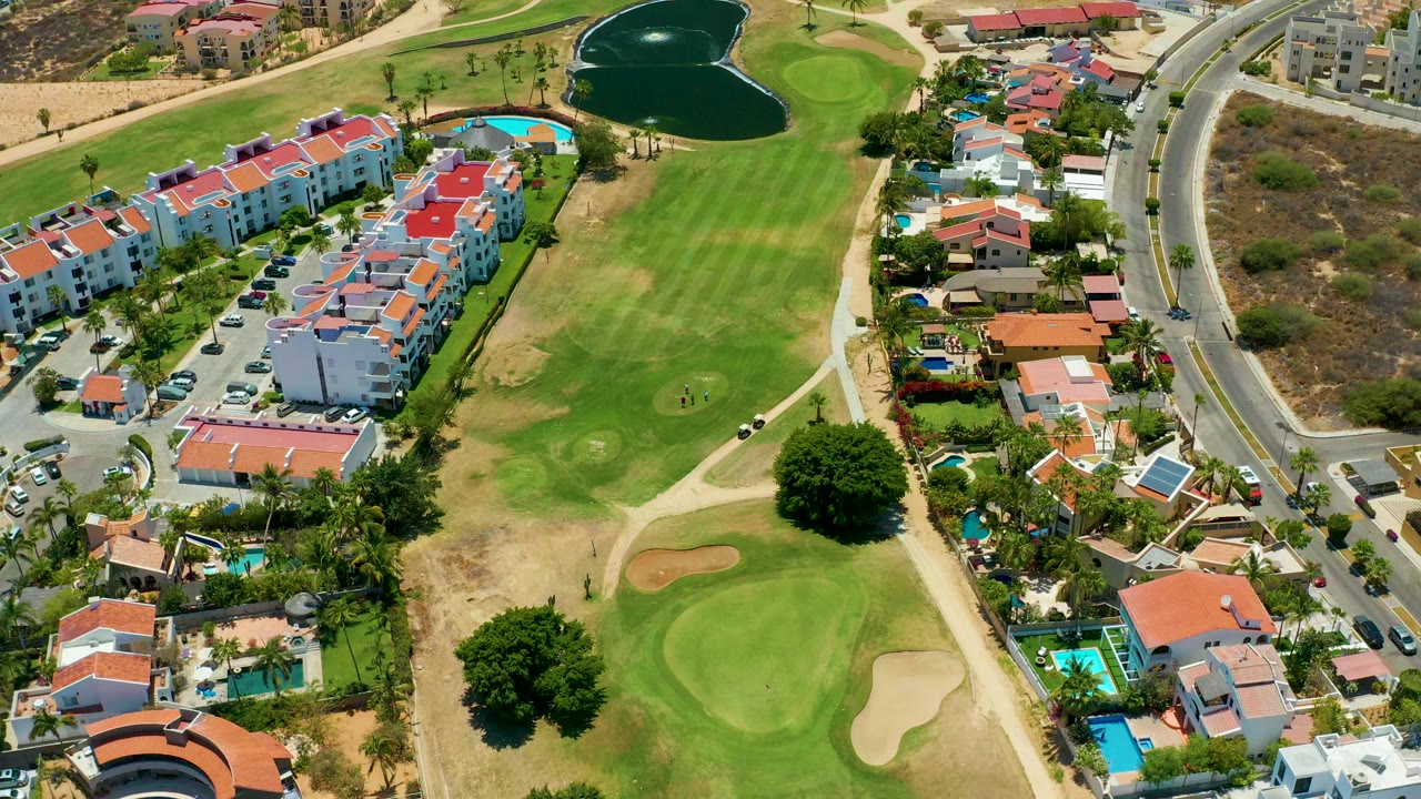 Opening aerial frame over San Jose Del Cabo with green golf fairways, red-roof residential buildings, blue pools, a dark pond, curving roads, and no visible people.