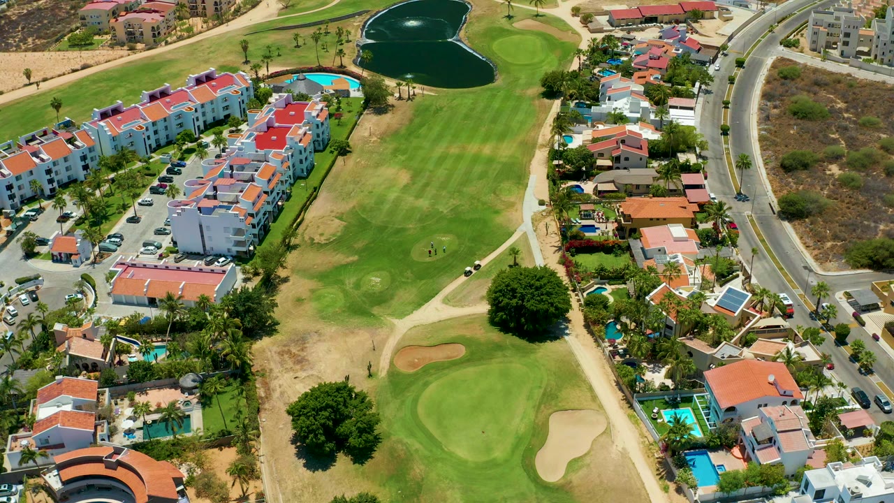 San Jose Del Cabo aerial frame with the fairway centered, sand traps near the lower green, residential buildings and pools on the left, roadway on the right, and no visible people.