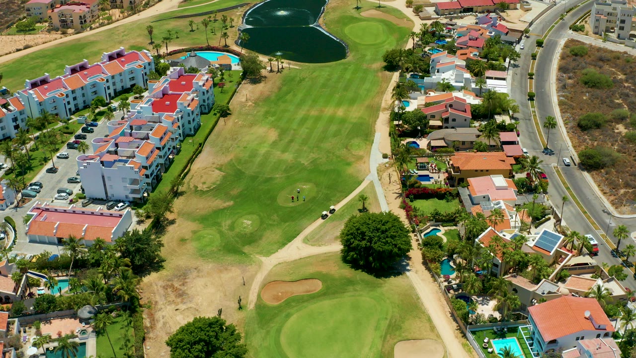 Opening aerial frame over a San Jose Del Cabo golf course with green fairways, sand traps, red-roof housing, a curved road, a pond, and no visible people.