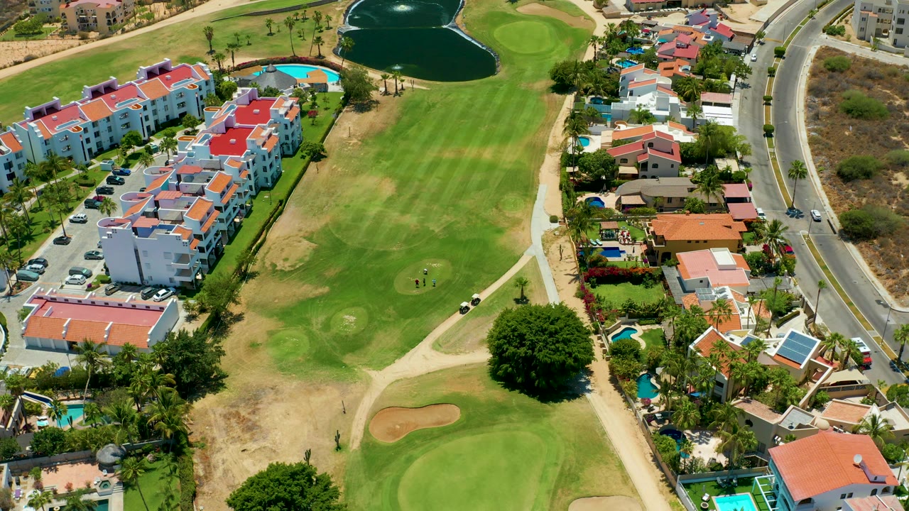 Third frame holds above the San Jose Del Cabo golf course with the dark pond near the top, red-roof buildings at left, houses at right, cars on the road, and no visible people.