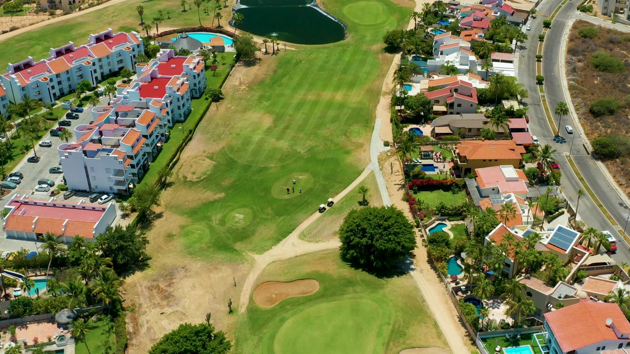 Drone view of fairways, sand traps, red-roof residences, pools, and a curved roadside.