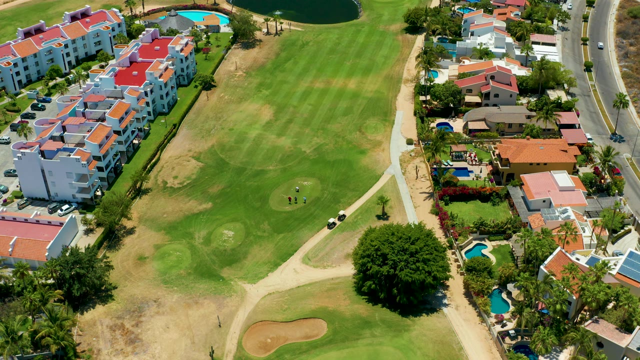 High downward frame over the San Jose del Cabo course with the fairway filling the center, the road along the right edge, homes and small pools beside it, and golfers still visible near the green.