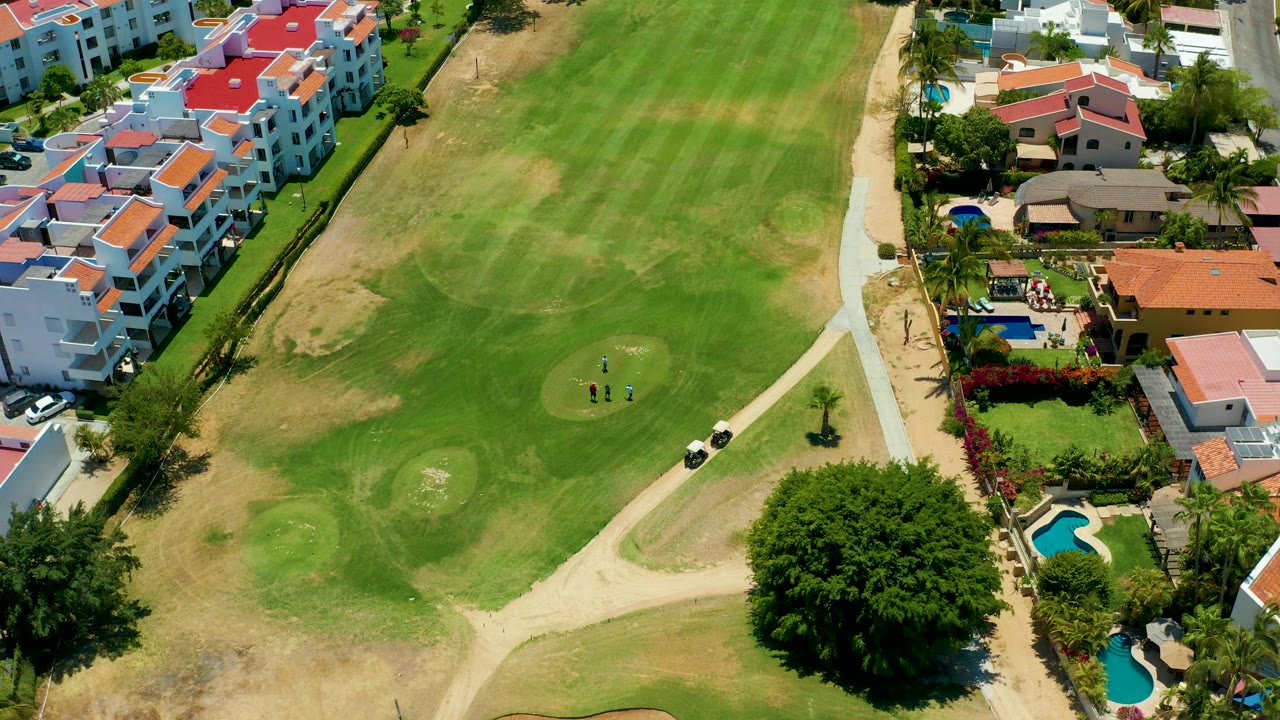 Overhead view in San Jose Del Cabo showing golfers on a green, a golf cart near a pale path, dry grass patches, white residences, red roofs, pools, and trees.
