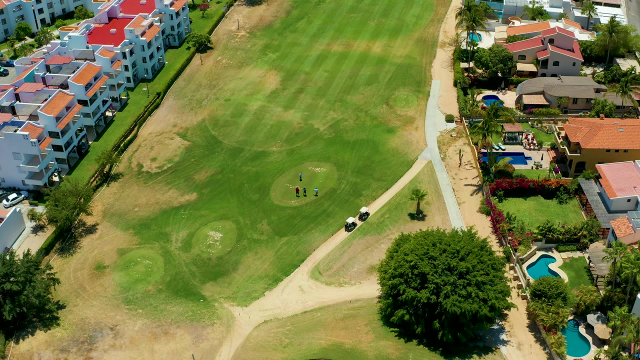 Overhead San Jose Del Cabo golf course view with golfers still visible on the green, a golf cart by the path, striped fairway grass, tan worn areas, villas, pools, and garden trees.