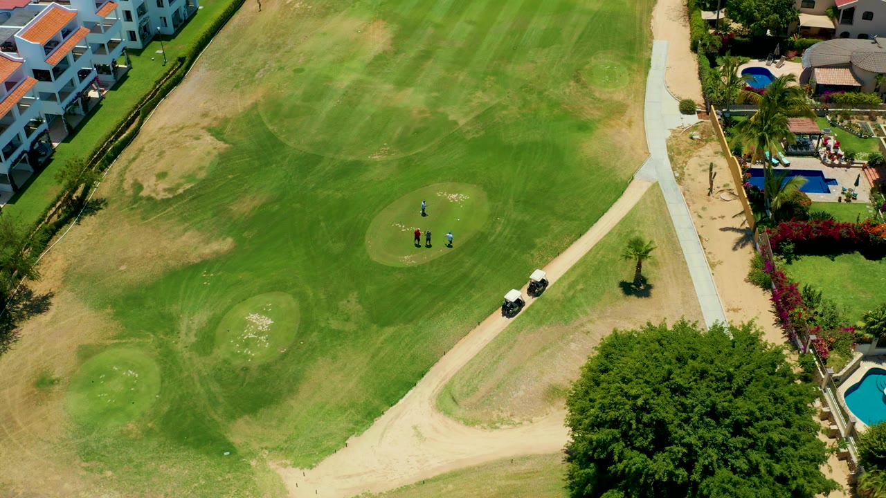Drone view of golfers on a green with carts beside a path and homes nearby.