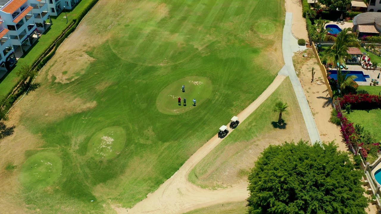 San Jose del Cabo aerial frame with a broad fairway, visible golfers near the green, carts by the path, patchy brown turf, sand traps, palms, homes, and pools in bright sun.