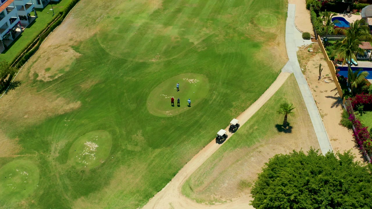 Aerial frame from San Jose Del Cabo showing small golfers on a green, two golf carts beside a pale path, sand traps, trees, buildings, and a pool at the edge of the course.