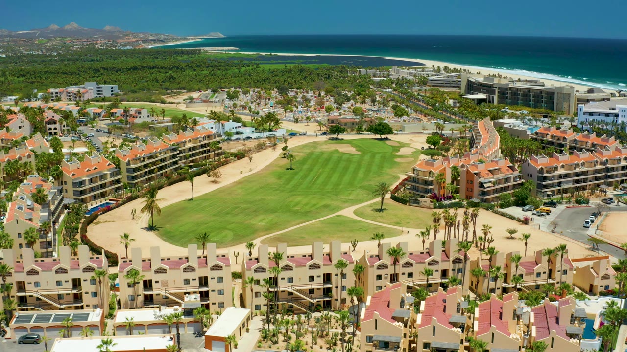 San Jose Del Cabo resort roofs line the lower frame while a manicured golf course, sandy beach, blue ocean, distant hills, and no visible people fill the view.