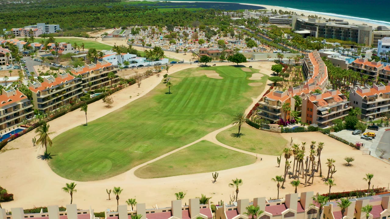 Aerial frame over San Jose del Cabo showing a green golf fairway, sand paths, resort buildings, palms, distant beach and ocean, with no visible people.