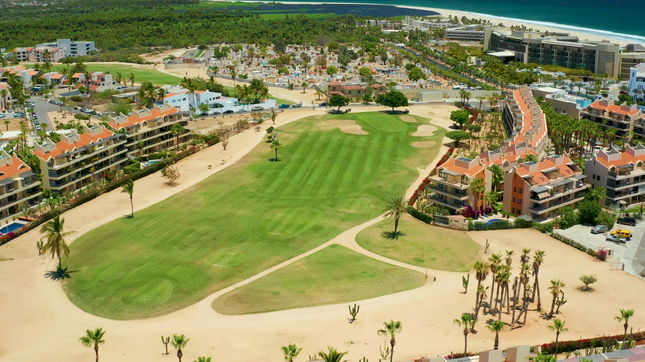 Aerial San Jose del Cabo frame showing the golf green and fairway, palm clusters, resort apartments with terracotta roofs, a sandy shoreline, and blue ocean, with no visible people.