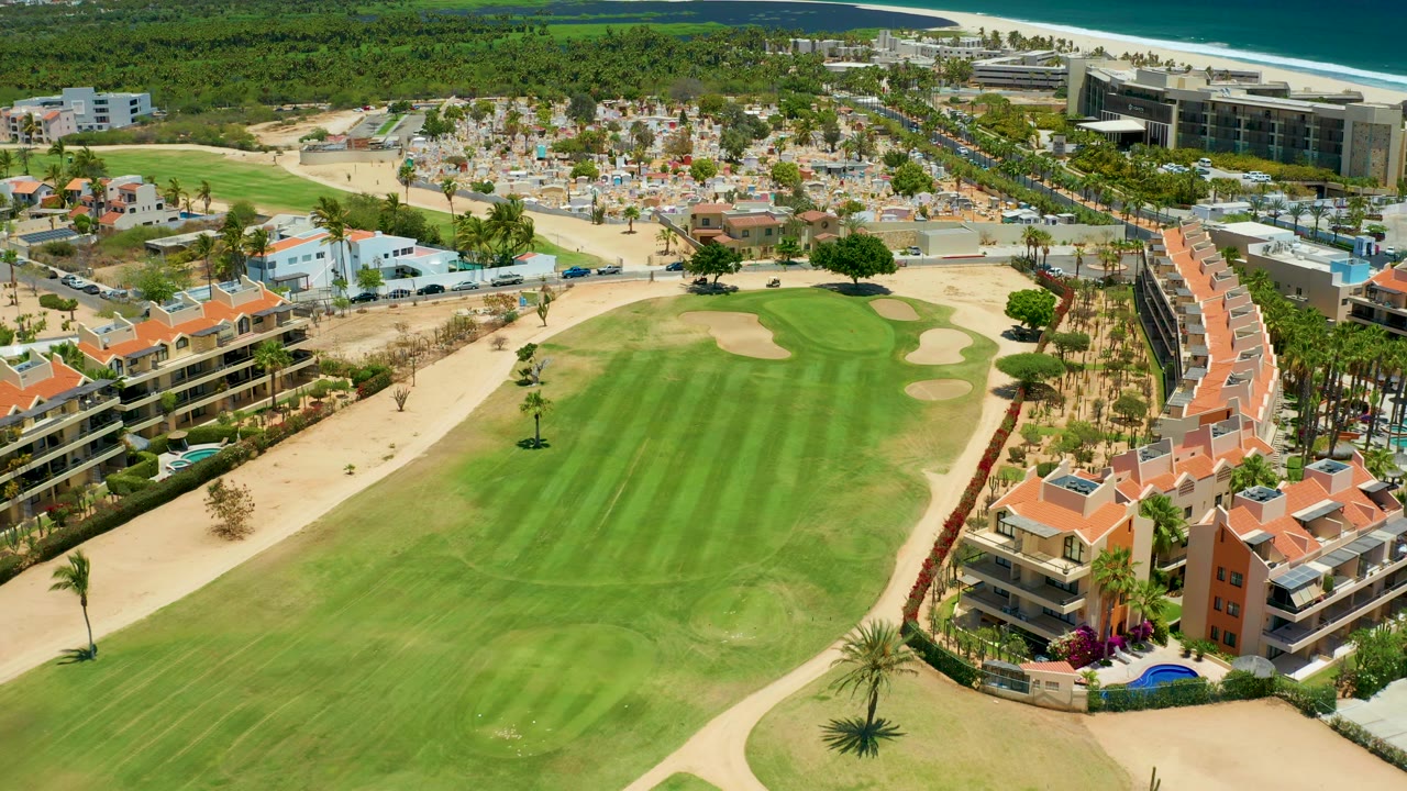 Drone view in San Jose Del Cabo looking across a green fairway and sand traps toward red-roof homes, palms, parking areas, beach, ocean, and no visible people.