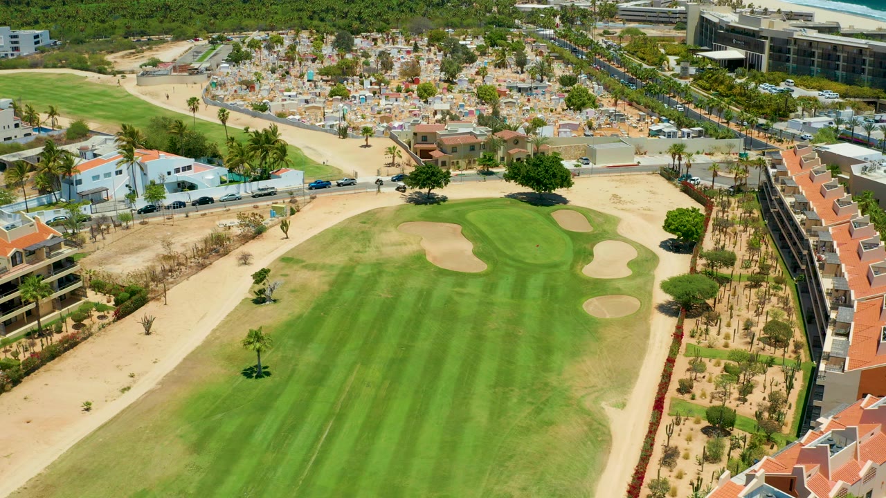 Aerial frame over a San Jose del Cabo golf fairway with sand traps, dry landscaped edges, nearby homes, cars on roads, and no visible people.