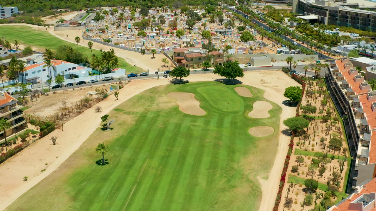The San Jose del Cabo aerial view shows a bright golf green with multiple sand traps, a curved road with vehicles, dry terrain, homes, and no visible people.