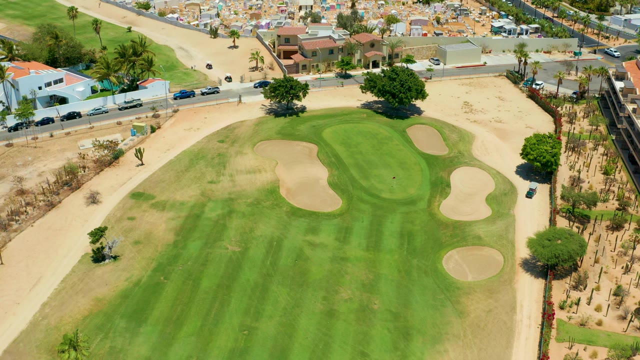 Aerial frame over a San Jose del Cabo golf course green with several tan sand traps, a road with cars, nearby homes, palm trees, sandy terrain, and no visible people.