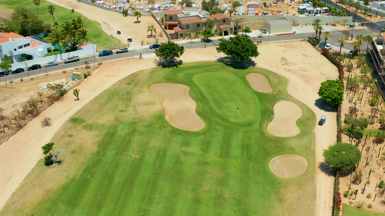 Aerial San Jose del Cabo frame with curved sand traps across the golf green, a road and parked or moving cars at the top, trees casting small shadows, sandy ground, and no visible people.