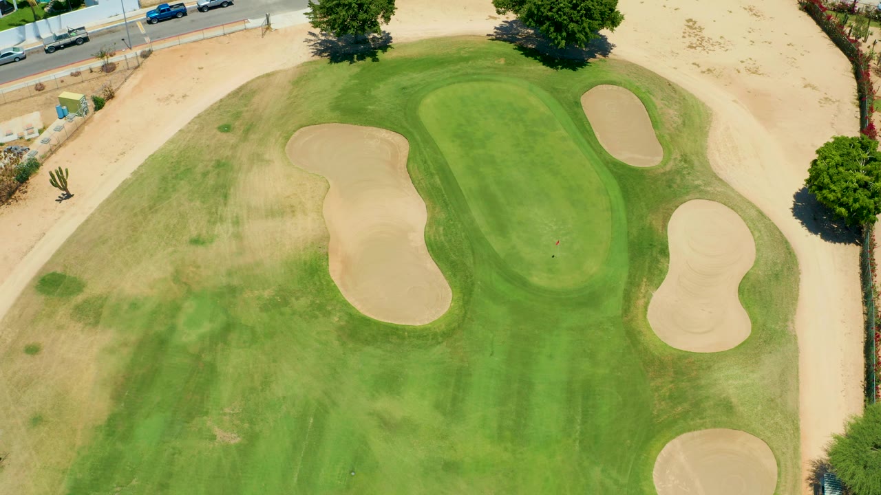 The aerial frame looks down on a San Jose del Cabo golf green with curved mowing patterns, pale bunkers, a sandy perimeter, a few roadside cars, and no visible people.