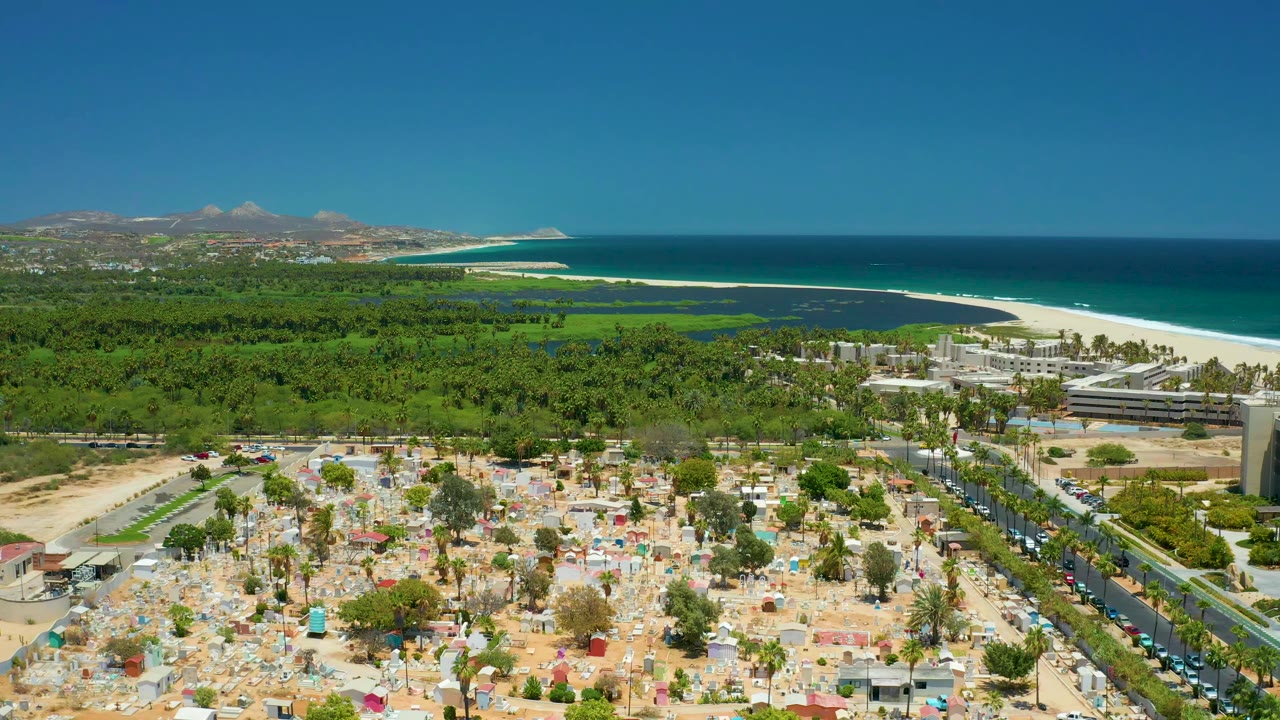 Opening aerial frame over San Jose del Cabo with a sandy foreground area, roadside cars at right, green tree canopy, beach, turquoise ocean, distant hills, clear sky, and no visible people.