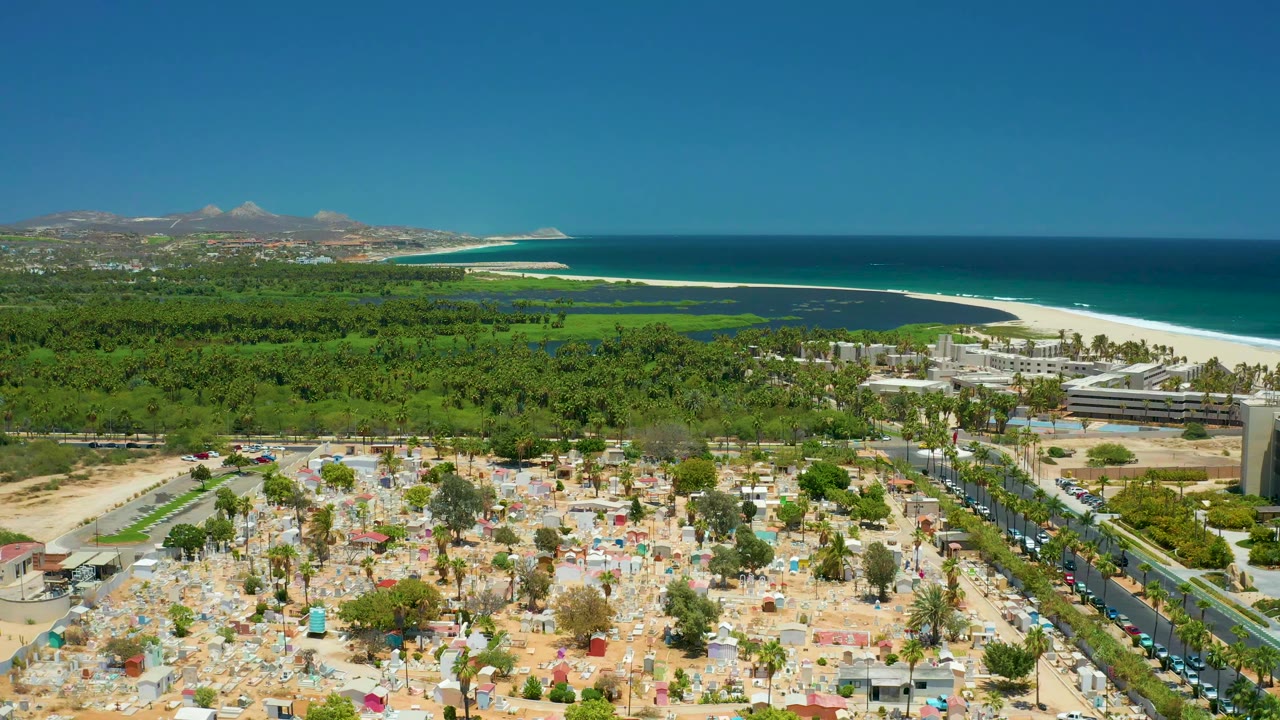 Aerial coastal view with sandy foreground, cars on a road, trees, white beach, and blue ocean.
