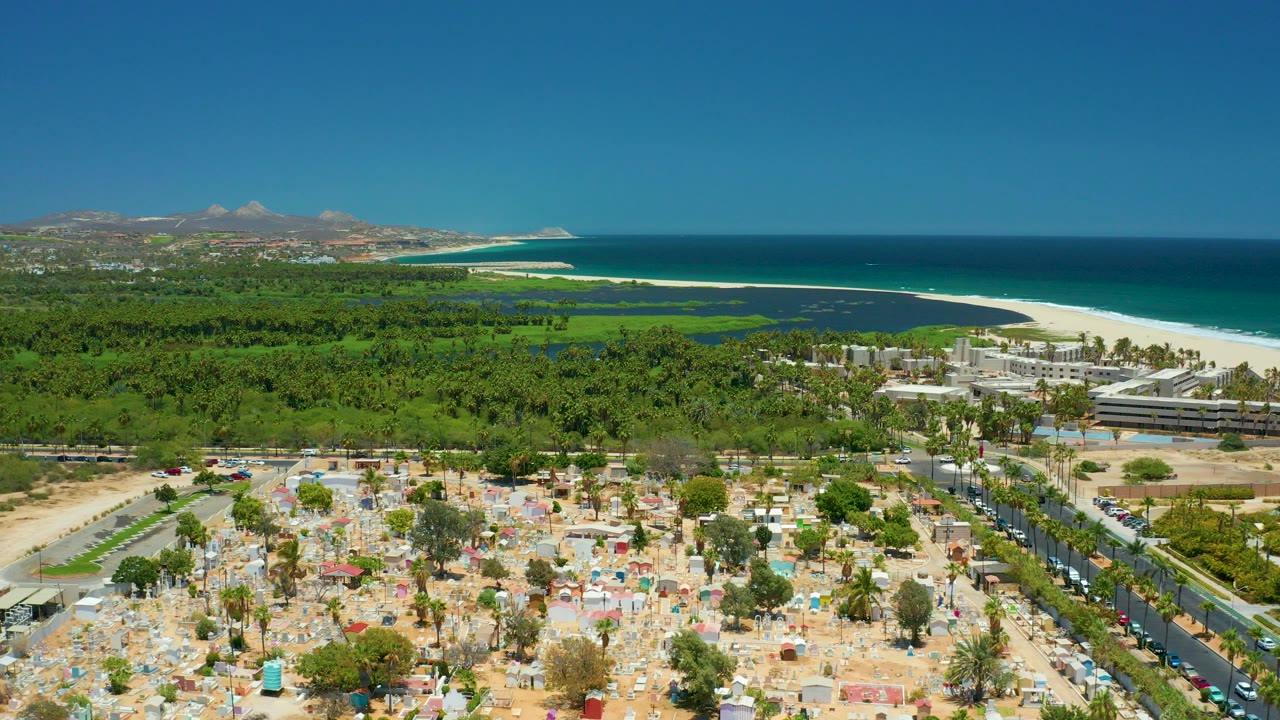 Aerial view in San Jose Del Cabo showing a sandy beach parking area with umbrellas, a coastal road, dense green trees, resort buildings, blue ocean, distant hills, and no visible people.