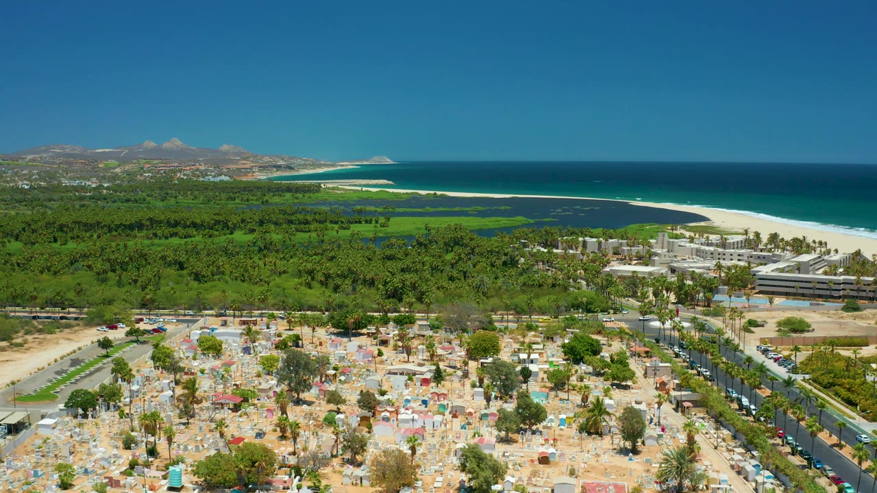 Drone-style view in San Jose Del Cabo with clustered umbrellas and parked beach area below, a road lined with palms and cars, a green belt, resort buildings, turquoise ocean, hills, and no visible people.