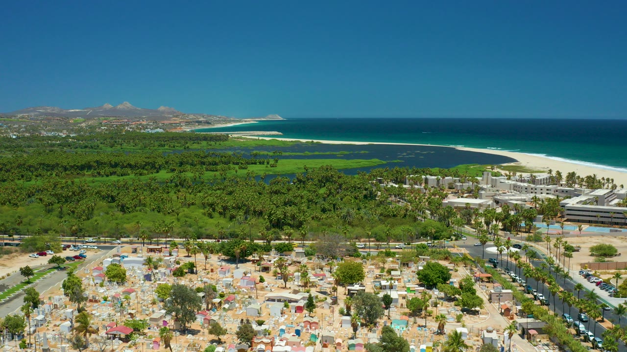 Wide aerial frame over San Jose del Cabo with a sandy parking area and umbrellas in front, green palms and a white beach beyond, ocean on the right, distant hills, and no visible people.