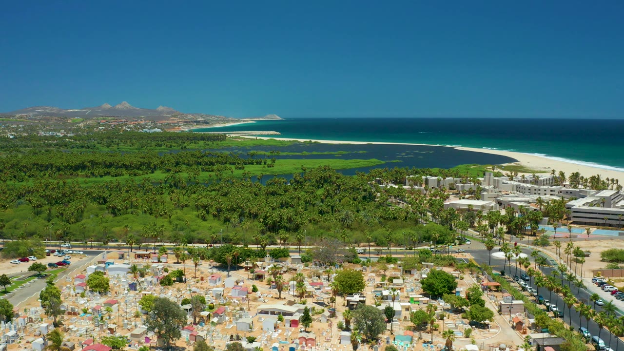 Aerial coastal view with foreground parking, palms, road, beach, ocean, and hills.