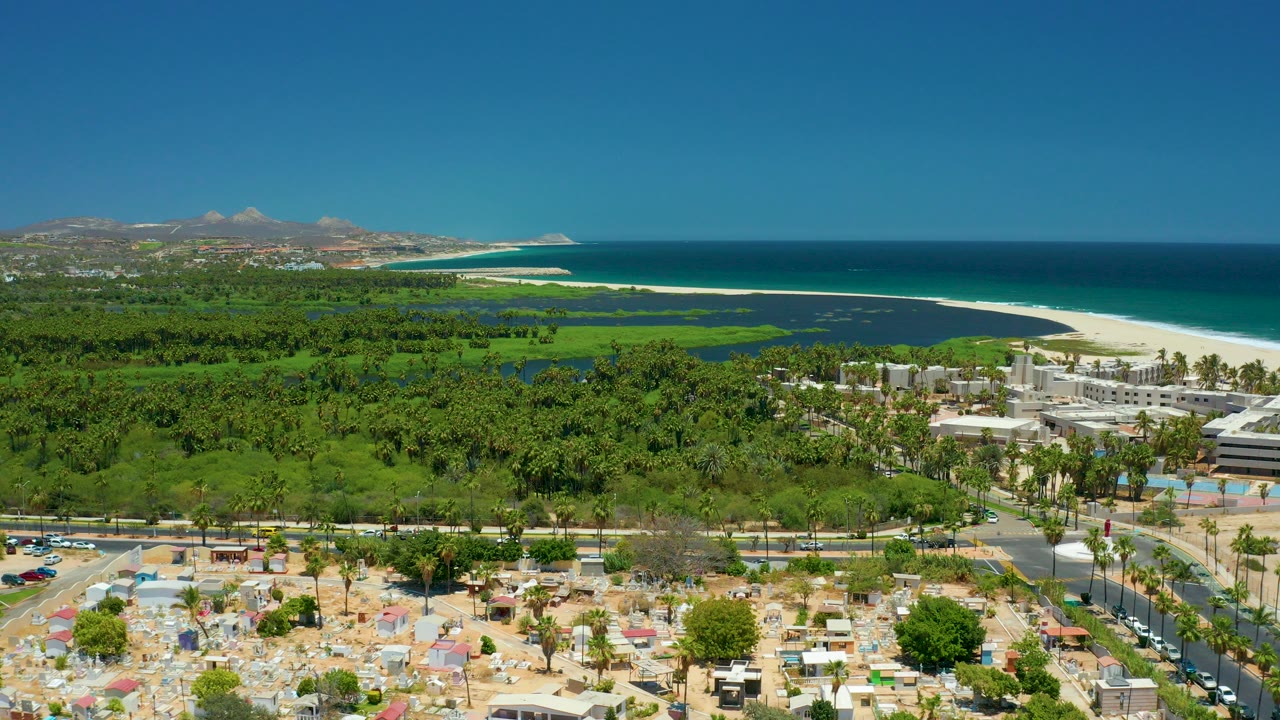 Opening San Jose del Cabo aerial frame with white beach and blue ocean behind a green palm area, foreground road, parked cars, umbrellas, and tiny visible people.