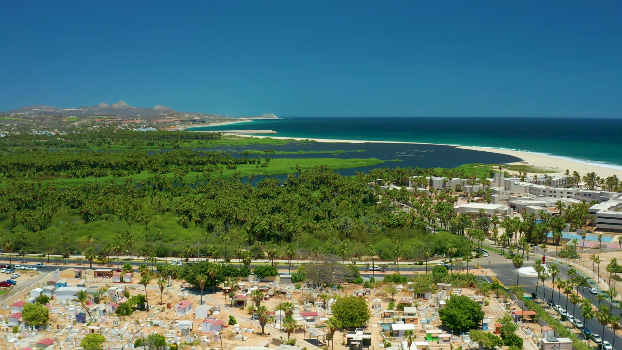 San Jose del Cabo frame at 0:02 with a broad green midground, resort-like buildings near the shoreline, foreground parking activity, and the ocean horizon under clear sky.